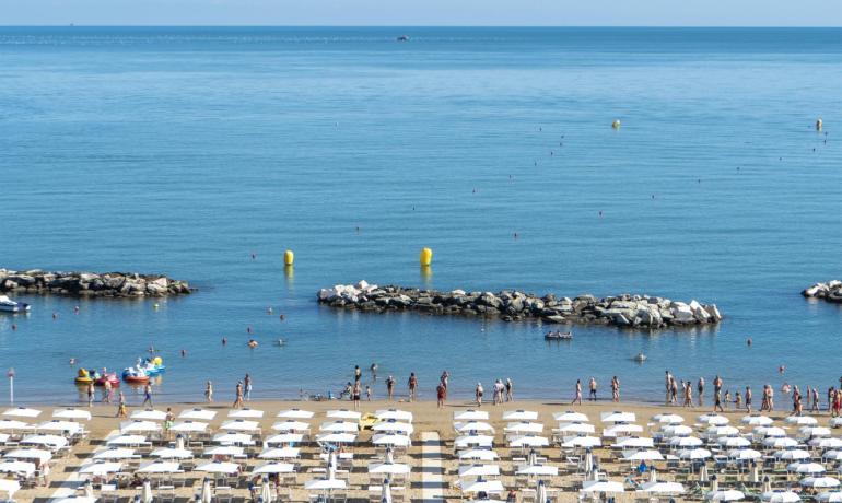 Spiaggia affollata, ombrelloni bianchi, mare calmo, cielo azzurro, giornata estiva perfetta.
