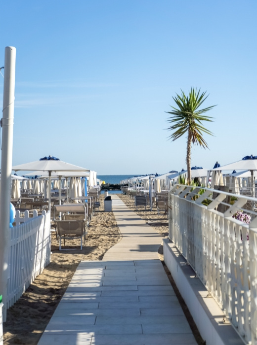 Sentiero sabbioso tra ombrelloni bianchi, palme e mare azzurro, atmosfera rilassante e vacanziera.