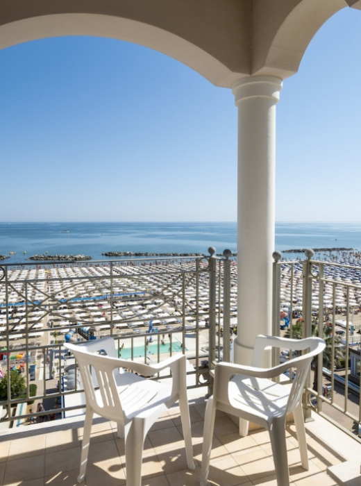 Vista mare da un balcone con sedie bianche e spiaggia affollata.