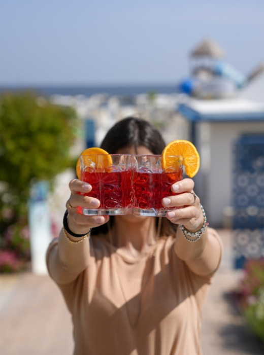 Persona offre due cocktail rossi con fette d'arancia in un ambiente estivo.