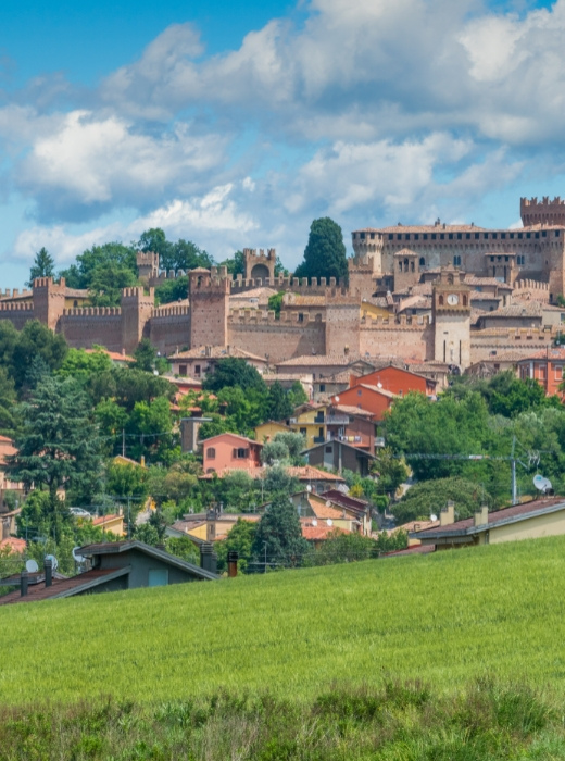 Un castello medievale domina colline verdi, case colorate e un cielo azzurro con nuvole bianche.
