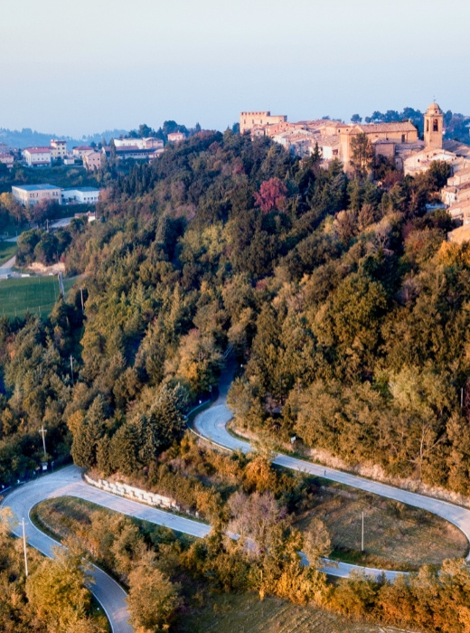 Strada serpeggiante tra colline boscose, conduce a un antico borgo affacciato sul panorama.