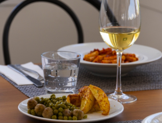 Piatto con piselli, patate, vino bianco; sfondo di pasta al pomodoro, atmosfera conviviale.
