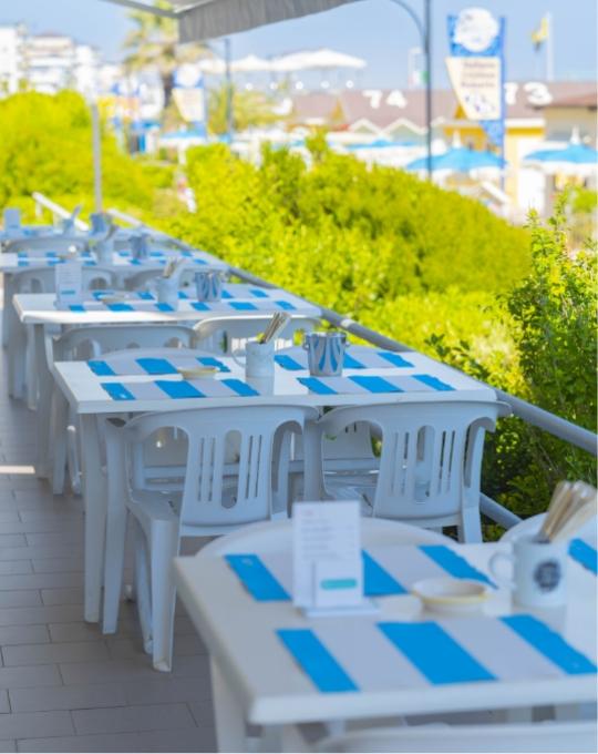 Tavoli bianchi all'aperto, tovagliette blu, atmosfera estiva e rilassante vicino al mare.