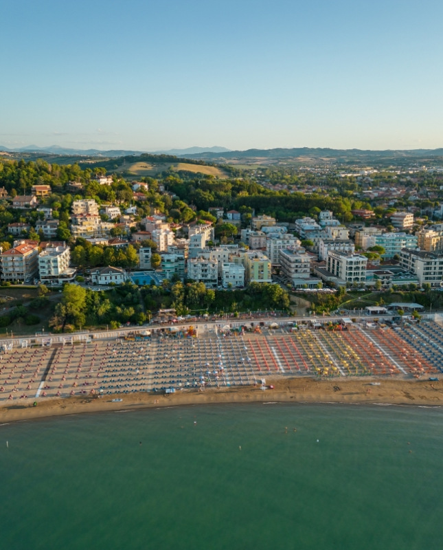 Spiaggia affollata, ombrelloni colorati, mare calmo, città collinare verdeggiante al tramonto.
