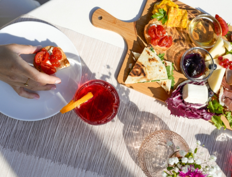 Colazione all'aperto: bruschette, formaggi e cocktail rosso, sole e atmosfera conviviale.