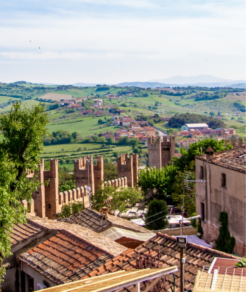 Paesaggio collinare con mura storiche, tetti antichi e verde lussureggiante sotto un cielo azzurro.