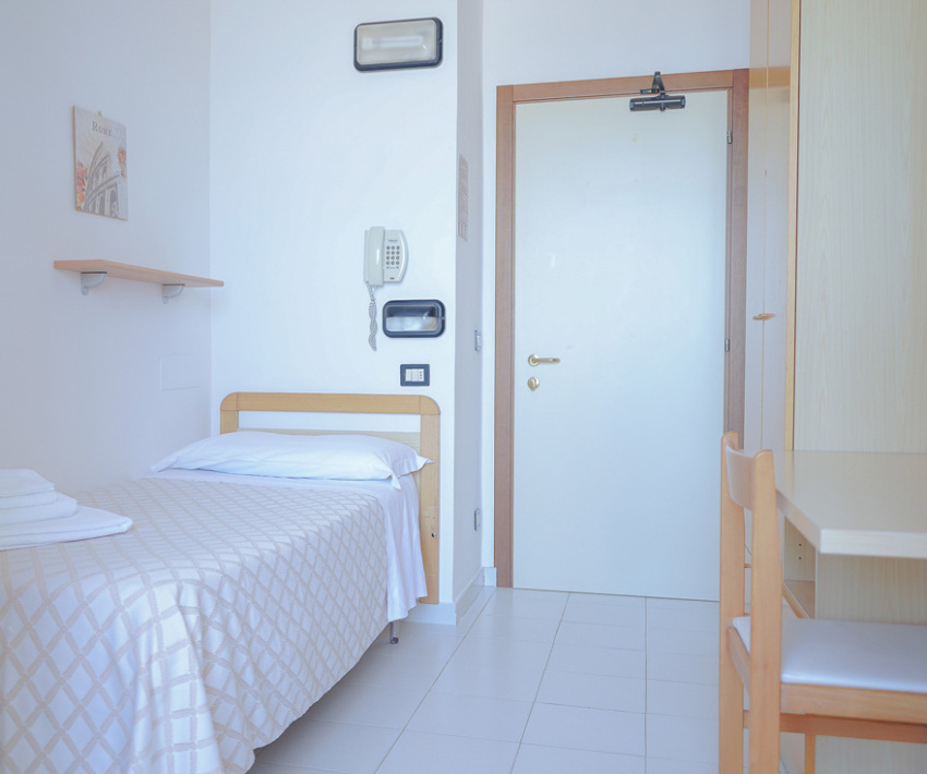 Camera singola minimalista con letto, scrivania e pareti bianche.