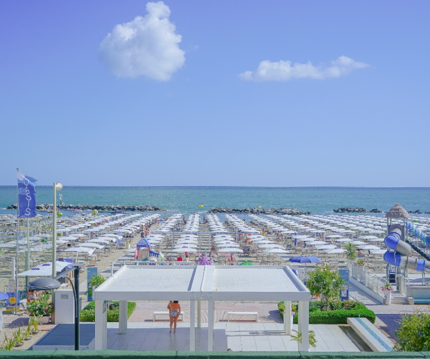 Spiaggia affollata, ombrelloni bianchi, mare blu, cielo sereno, atmosfera estiva e rilassante.